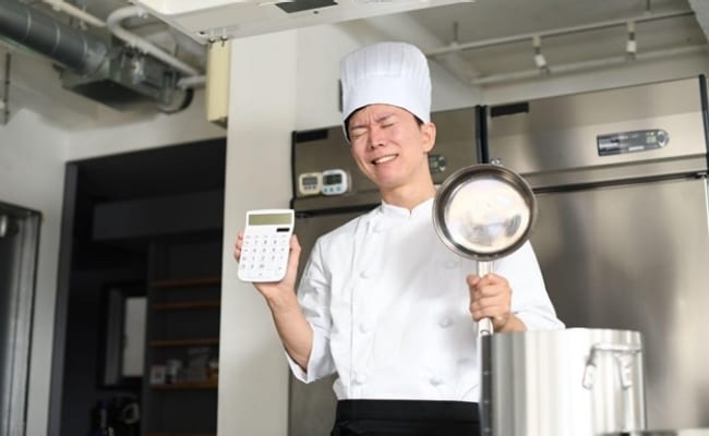 Seperti yang terlihat pada foto, ada chef yang sedang stres sambil memegang pan dan kalkulator. 