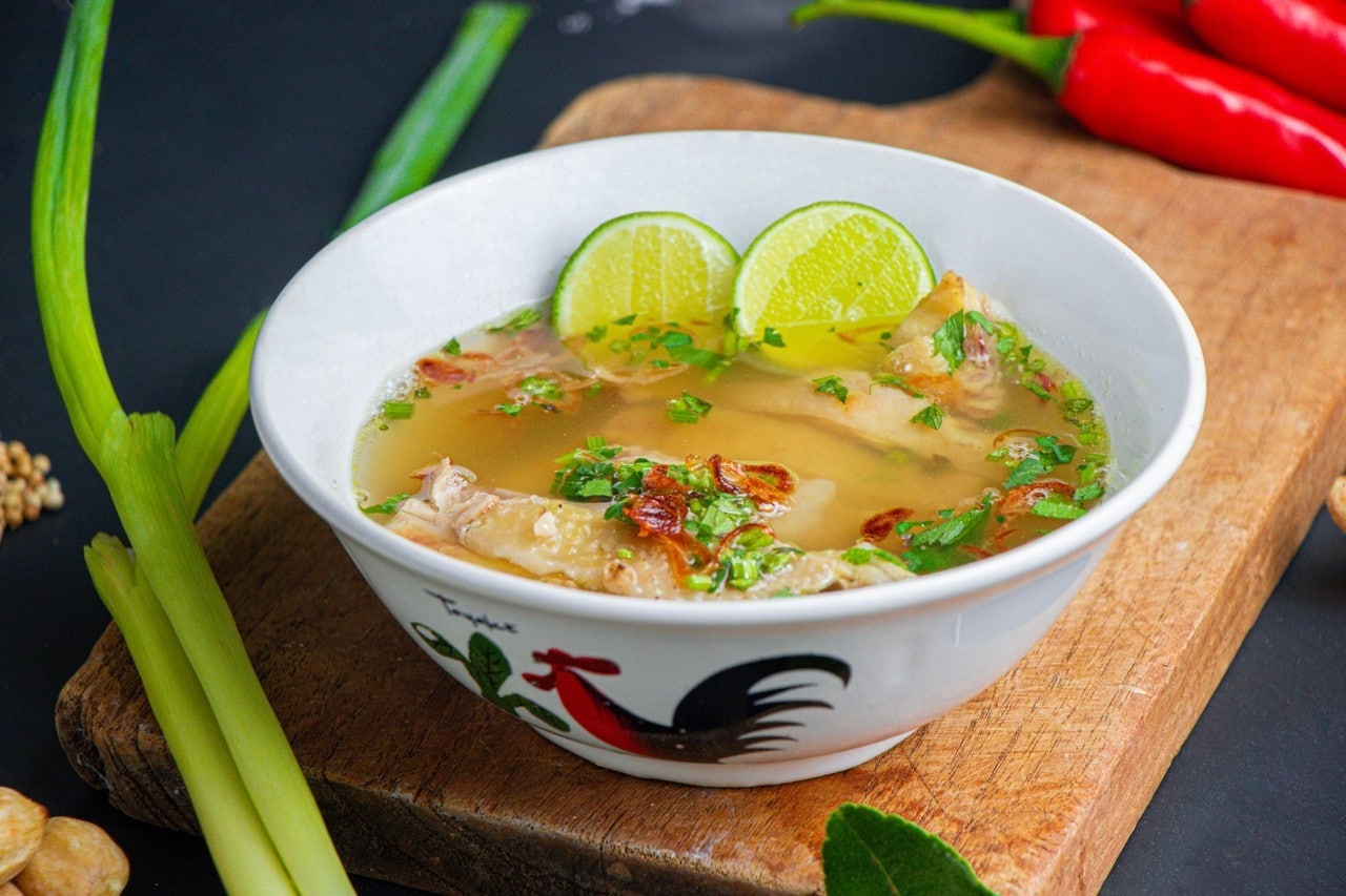 Mangkuk berisi sup ayam pedas di atas talenan, dikelilingi bahan segar.