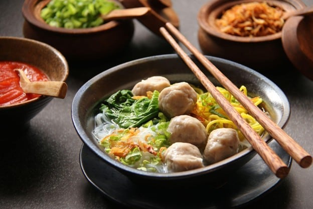 Seperti yang terlihat pada gambar, semangkuk bakso kuah dengan saus sambal, bawang goreng, dan seledri