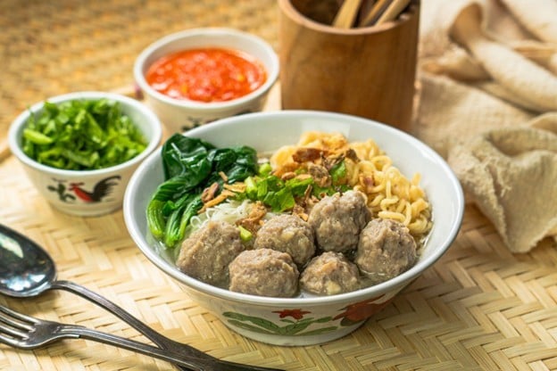 Seperti yang terlihat pada gambar, semangkuk bakso dikelilingi saus, sendok garpu, dan potongan seledri