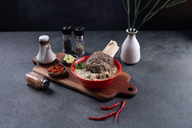 Seperti yang terlihat pada gambar, seporsi bakso iga dalam mangkuk merah di atas talenan kayu
