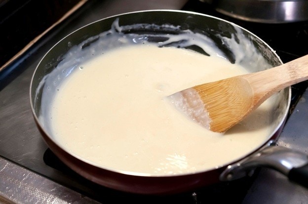 Seperti yang terlihat pada foto, ada saus bechamel berwarna putih creamy di dalam wajan dengan sendok kayu.