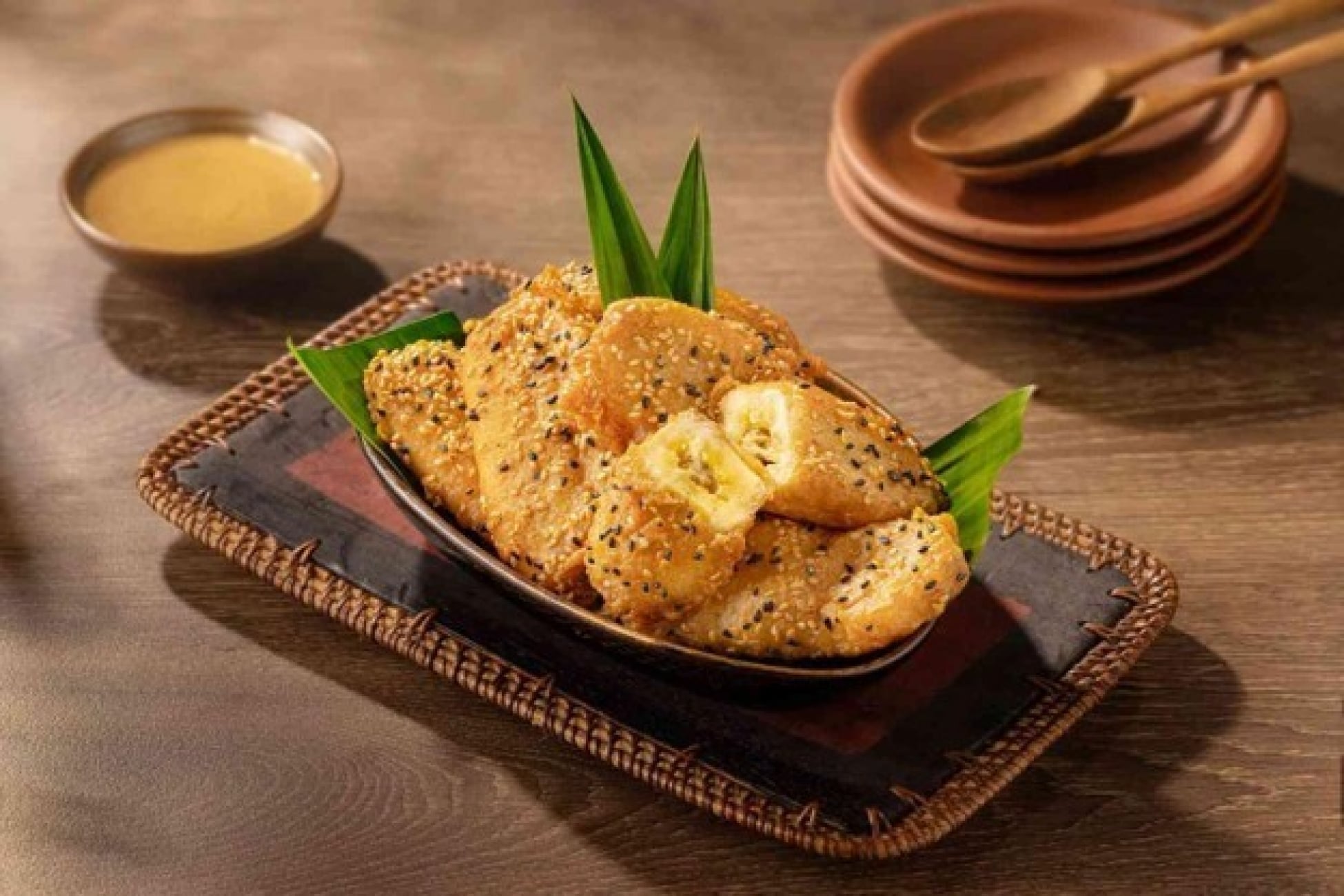 Pisang goreng bertabur wijen disajikan di atas daun pandan dengan saus pendamping.