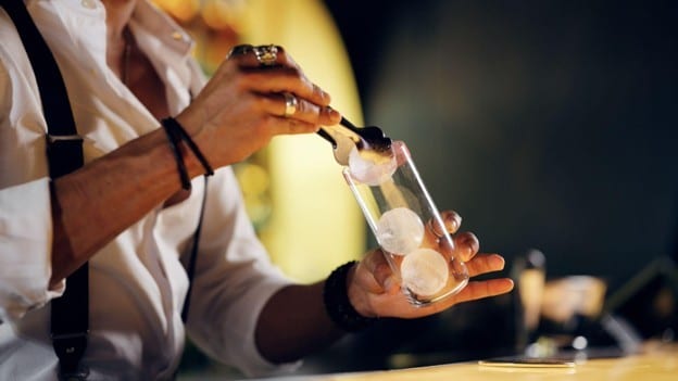 Seperti yang terlihat pada foto, bartender sedang memasukkan es batu ke dalam gelas.